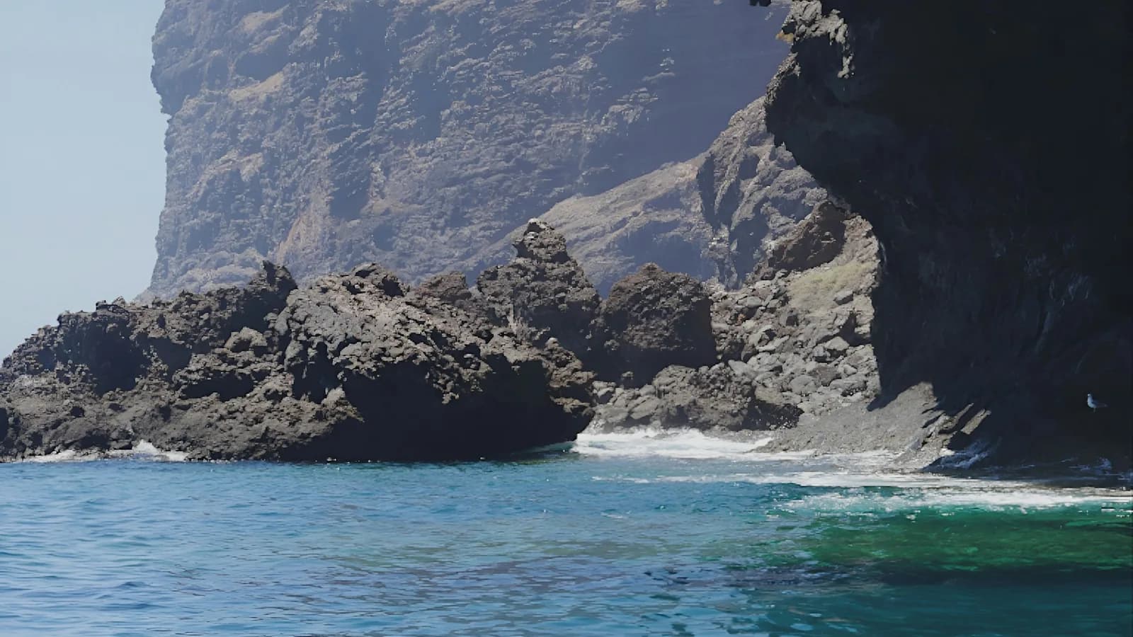 Steil abfallende Vulkanklippe über dunklem Lavagestein und türkisem Wasser